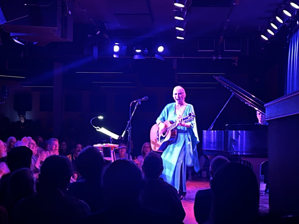 Judy Collins in a blue dress and short hair faces the crowd at The Ark